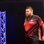 Michael Smith walking during PDC Europe Darts Matchplay with a black background, lit up by two colourful light pillars.