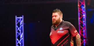 Michael Smith walking during PDC Europe Darts Matchplay with a black background, lit up by two colourful light pillars.