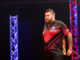 Michael Smith walking during PDC Europe Darts Matchplay with a black background, lit up by two colourful light pillars.